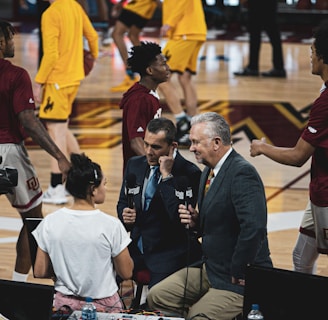 A group of individuals, likely basketball players, is on a gymnasium court. Some are wearing yellow athletic outfits, and others are in burgundy athletic gear. Two men in suits are speaking into microphones while a woman stands nearby wearing a white shirt. A camera setup is positioned to capture the scene, indicating a broadcast or interview setting.