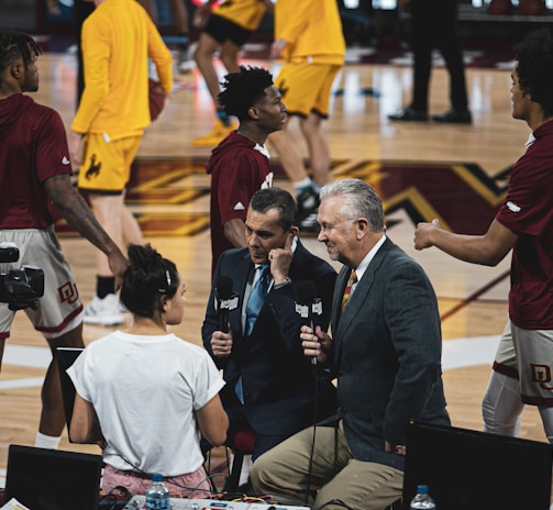 A group of individuals, likely basketball players, is on a gymnasium court. Some are wearing yellow athletic outfits, and others are in burgundy athletic gear. Two men in suits are speaking into microphones while a woman stands nearby wearing a white shirt. A camera setup is positioned to capture the scene, indicating a broadcast or interview setting.