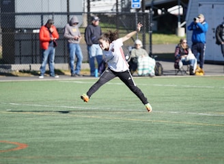 A person is mid-air on a sports field, appearing to be involved in an athletic activity. Several spectators are on the sidelines, some seated and others standing, with one individual capturing the event with a camera. The background includes a fence and various casual observers, some of whom are wearing winter clothing.