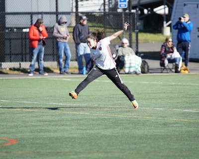 A person is mid-air on a sports field, appearing to be involved in an athletic activity. Several spectators are on the sidelines, some seated and others standing, with one individual capturing the event with a camera. The background includes a fence and various casual observers, some of whom are wearing winter clothing.