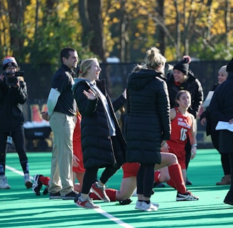 A group of people on a sports field, including several athletes in red uniforms and coaches wearing black coats. Some individuals are kneeling, while others are standing, engaged in animated conversation. A photographer or videographer is capturing the scene beside them. The background features a fence and autumn trees with yellow leaves.