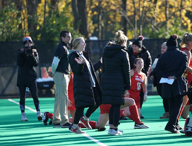 A group of people on a sports field, including several athletes in red uniforms and coaches wearing black coats. Some individuals are kneeling, while others are standing, engaged in animated conversation. A photographer or videographer is capturing the scene beside them. The background features a fence and autumn trees with yellow leaves.