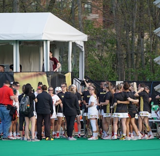 A group of athletes in sports uniforms is gathered on what appears to be a lacrosse field. They are standing in two groups, some wearing white uniforms and some wearing dark uniforms. There is a tent with people inside, possibly officials or commentators, overlooking the field in the background. Extensive greenery and trees are visible behind the tent.