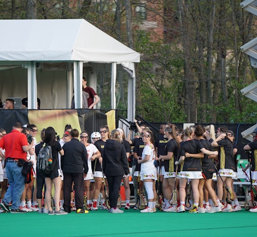 A group of athletes in sports uniforms is gathered on what appears to be a lacrosse field. They are standing in two groups, some wearing white uniforms and some wearing dark uniforms. There is a tent with people inside, possibly officials or commentators, overlooking the field in the background. Extensive greenery and trees are visible behind the tent.