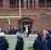 A group of people on a sports field, with some dressed in athletic gear, and others in casual clothing. A brick building labeled 'Gosset Football Team House' is in the background. One of the athletes appears to be aided by others, suggesting a possible injury.