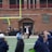 A group of people on a sports field, with some dressed in athletic gear, and others in casual clothing. A brick building labeled 'Gosset Football Team House' is in the background. One of the athletes appears to be aided by others, suggesting a possible injury.
