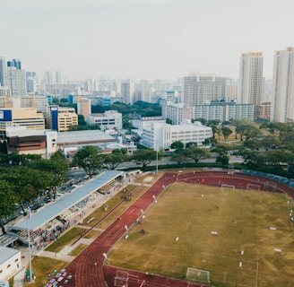 An aerial view of a sports complex with a running track surrounding a grassy field, set against the background of a cityscape. The city's skyline features a mix of tall residential buildings and shorter commercial structures. There are trees and green areas surrounding the sports facility.