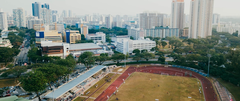 An aerial view of a sports complex with a running track surrounding a grassy field, set against the background of a cityscape. The city's skyline features a mix of tall residential buildings and shorter commercial structures. There are trees and green areas surrounding the sports facility.