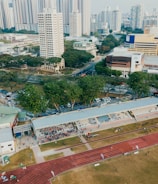 An aerial view of an urban area featuring a sports complex with a track field and a grandstand. Surrounding the sports area are numerous high-rise buildings, greenery, and roads with vehicles. The image captures people engaging in activities on the field.