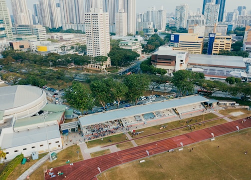 An aerial view of an urban area featuring a sports complex with a track field and a grandstand. Surrounding the sports area are numerous high-rise buildings, greenery, and roads with vehicles. The image captures people engaging in activities on the field.