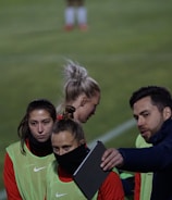 A group of individuals on a sports field are wearing green vests over red attire. One person is holding a tablet and seems to be discussing or showing something to the others. The atmosphere suggests a sports event, likely soccer, indicated by their clothing and the setting. The background is a grassy field.