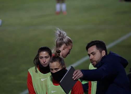 A group of individuals on a sports field are wearing green vests over red attire. One person is holding a tablet and seems to be discussing or showing something to the others. The atmosphere suggests a sports event, likely soccer, indicated by their clothing and the setting. The background is a grassy field.