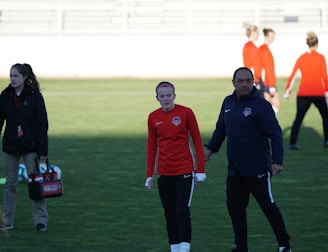 A group of people are gathered on a sports field. Some individuals are dressed in athletic gear, including a woman in a red sports jersey and a man beside her in a blue jacket, appearing to be in conversation. Additional people in similar athletic clothing are visible in the background.