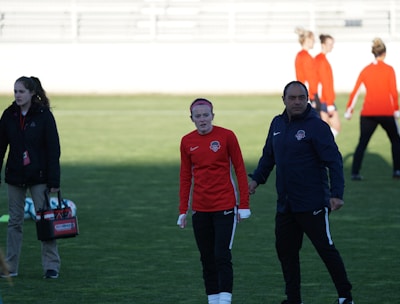 A group of people are gathered on a sports field. Some individuals are dressed in athletic gear, including a woman in a red sports jersey and a man beside her in a blue jacket, appearing to be in conversation. Additional people in similar athletic clothing are visible in the background.