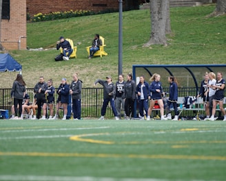 A group of female athletes, some in sports uniforms and others in jackets, are gathered along the sideline of a sports field. They seem to be in a team environment, possibly preparing for or observing a game. In the background, there are a few people sitting on yellow chairs on a grassy incline, and a building with a brick facade.