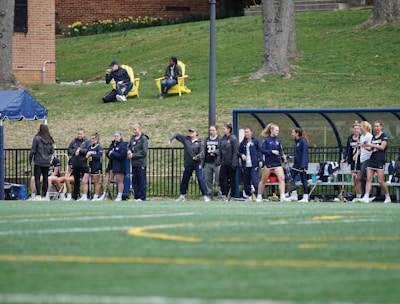 A group of female athletes, some in sports uniforms and others in jackets, are gathered along the sideline of a sports field. They seem to be in a team environment, possibly preparing for or observing a game. In the background, there are a few people sitting on yellow chairs on a grassy incline, and a building with a brick facade.