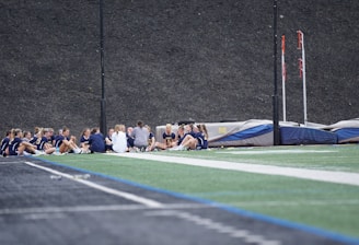 A group of people, likely a sports team, is sitting together on a field. They are wearing matching athletic uniforms, suggesting team unity, and seem to be engaged in a discussion or meeting. In the background, there is sports equipment and a high fence.