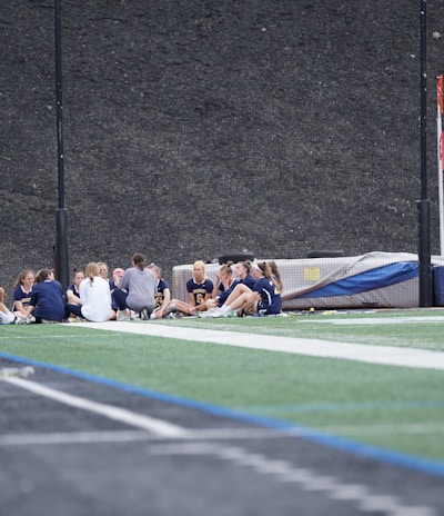 A group of people, likely a sports team, is sitting together on a field. They are wearing matching athletic uniforms, suggesting team unity, and seem to be engaged in a discussion or meeting. In the background, there is sports equipment and a high fence.