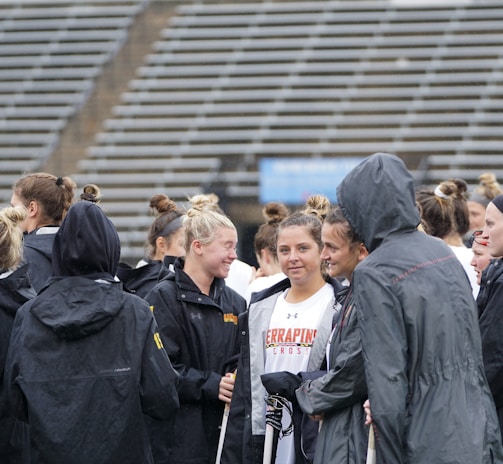 A group of people, likely athletes, are gathered together on a sports field. They are wearing black jackets with some individuals dressed in white sports uniforms underneath. A few people have their hair tied up in buns, and most are engaged in conversation. The background shows tiered stadium seats, suggesting the setting is an outdoor sports venue.