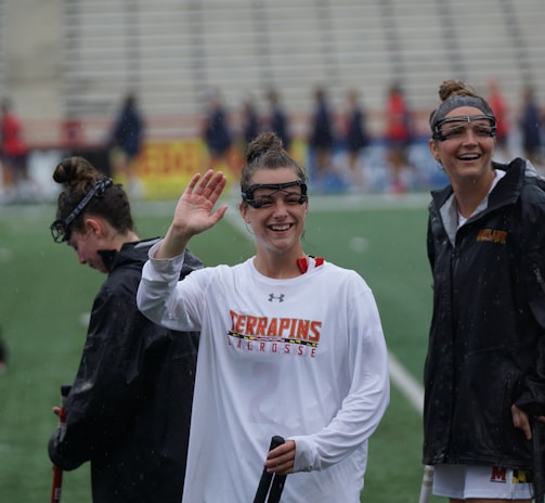 Several athletes in sports attire are on an outdoor sports field, interacting and smiling. One individual in the foreground waves with a friendly expression, while another stands beside her, also smiling. They wear protective goggles and gear associated with lacrosse. The background features indistinct figures in dark clothing against a backdrop of stadium seating.