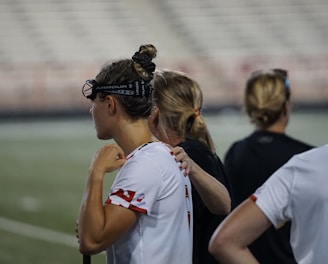 Several people wearing sports attire and protective eyewear are gathered on a sports field. They appear to be engaged in a discussion or briefing, possibly during a break from a game or practice. The background shows a blurred stadium seating, indicating an outdoor setting.