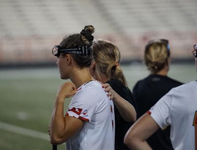 Several people wearing sports attire and protective eyewear are gathered on a sports field. They appear to be engaged in a discussion or briefing, possibly during a break from a game or practice. The background shows a blurred stadium seating, indicating an outdoor setting.
