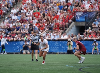 A sports event with two athletes engaged in a lacrosse game. One athlete in a white jersey is holding a lacrosse stick, appearing to prepare for a play. A referee in a striped uniform is nearby. The background features a large audience seated in a stadium, with many spectators wearing bright and casual clothing. Some photographers are also visible near the field.