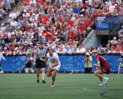 A sports event with two athletes engaged in a lacrosse game. One athlete in a white jersey is holding a lacrosse stick, appearing to prepare for a play. A referee in a striped uniform is nearby. The background features a large audience seated in a stadium, with many spectators wearing bright and casual clothing. Some photographers are also visible near the field.