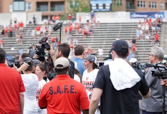 A lively gathering in an outdoor sports venue with a group of people, some wearing matching shirts labeled 'National Champions.' Several individuals are holding cameras and microphones, indicating a media presence. The background shows blurred spectators sitting on bleachers.