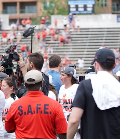 A lively gathering in an outdoor sports venue with a group of people, some wearing matching shirts labeled 'National Champions.' Several individuals are holding cameras and microphones, indicating a media presence. The background shows blurred spectators sitting on bleachers.