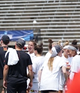 A group of athletes in white shirts with black and red accents are gathered together, engaged in conversation and smiling. They appear to be on a sports field with empty bleachers in the background. The setting is outdoors, and it seems sunny. Some individuals are wearing caps or headbands.