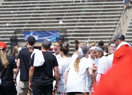 A group of athletes in white shirts with black and red accents are gathered together, engaged in conversation and smiling. They appear to be on a sports field with empty bleachers in the background. The setting is outdoors, and it seems sunny. Some individuals are wearing caps or headbands.