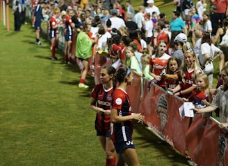 The image features a lively gathering of young soccer players and fans next to a field. Several female athletes in red and blue uniforms interact with an enthusiastic crowd standing behind a barrier. Some people in the crowd are holding objects, like pens and paper, possibly for autographs.