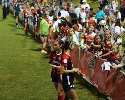 The image features a lively gathering of young soccer players and fans next to a field. Several female athletes in red and blue uniforms interact with an enthusiastic crowd standing behind a barrier. Some people in the crowd are holding objects, like pens and paper, possibly for autographs.
