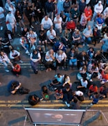 A large group of people gathered outdoors, watching a screen that is displaying a man. The crowd is diverse in age and appears interested and engaged, with many sitting closely together, some standing. Many are dressed casually, some in sports attire, and a few waving or holding flags. The event seems to have a communal, festive atmosphere.
