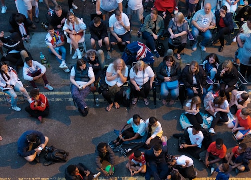 A large group of people gathered outdoors, watching a screen that is displaying a man. The crowd is diverse in age and appears interested and engaged, with many sitting closely together, some standing. Many are dressed casually, some in sports attire, and a few waving or holding flags. The event seems to have a communal, festive atmosphere.