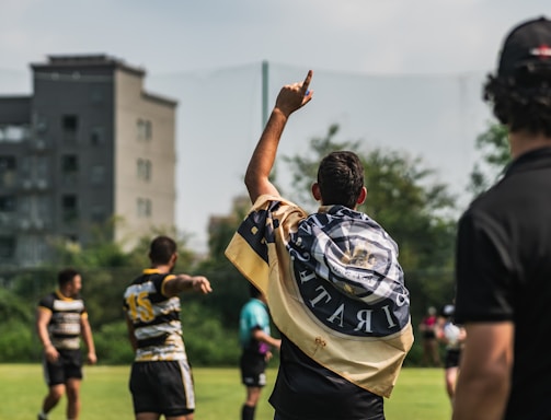 A group of athletes are on a grassy field engaged in a sports activity, likely rugby. One player is seen with a flag draped over their shoulders, pointing upwards. Other players and a referee or coach are visible, all wearing sports uniforms. In the background, there are trees and a building, suggesting an urban setting.